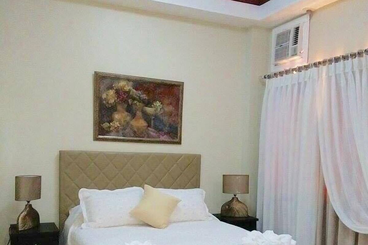 Une chambre à coucher bien rangée, avec un grand lit, deux tables de chevet avec des lampes, un tableau au mur et une fenêtre avec des rideaux.