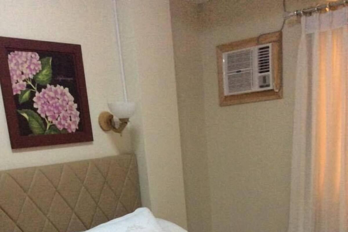 Un lit bien fait, avec une tête de lit matelassée, une œuvre d’art florale encadrée au mur et un petit appareil de climatisation placé au-dessus du lit.
