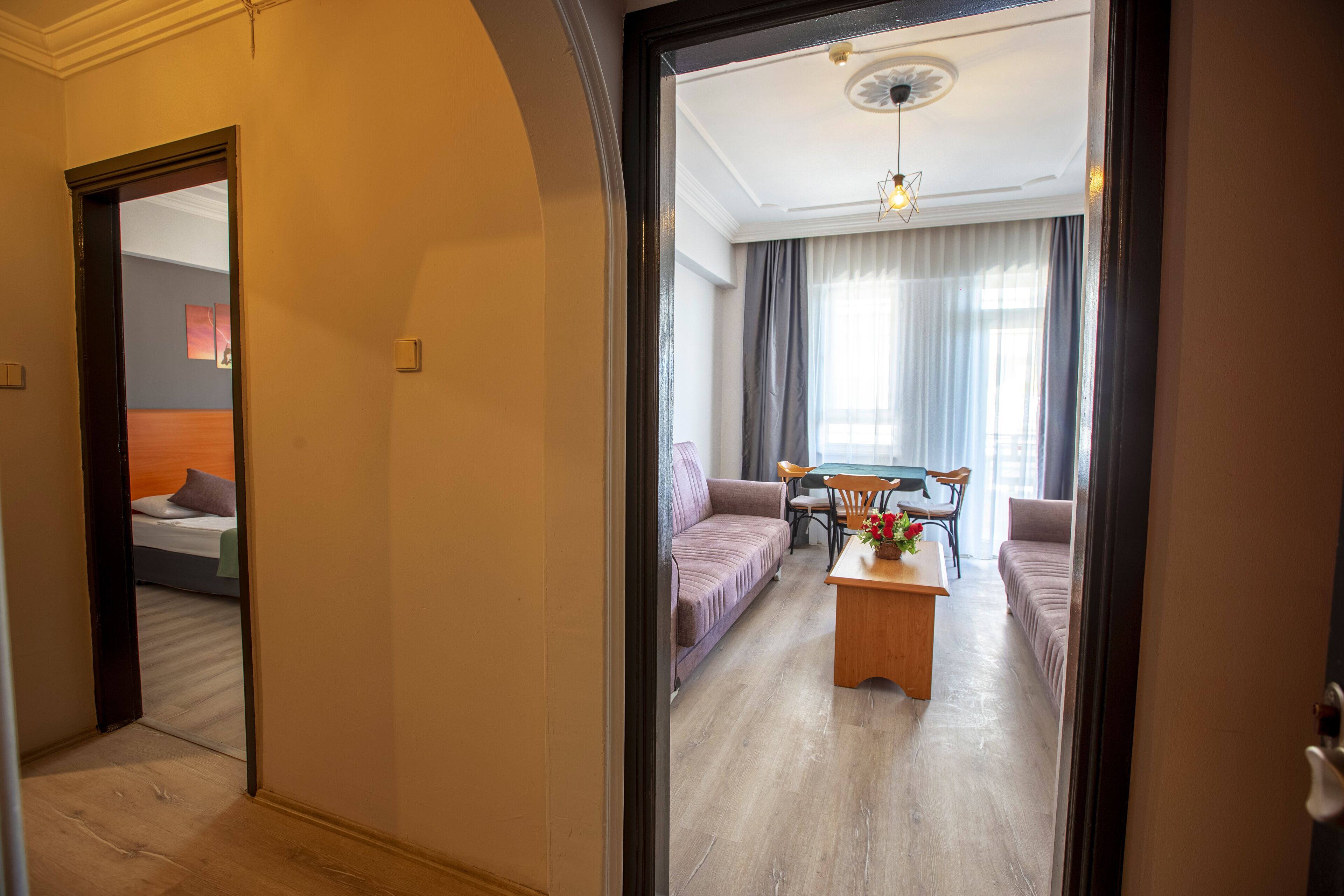A modern hotel room with a wooden floor, a sofa, a dining table with chairs, and a large window with curtains.