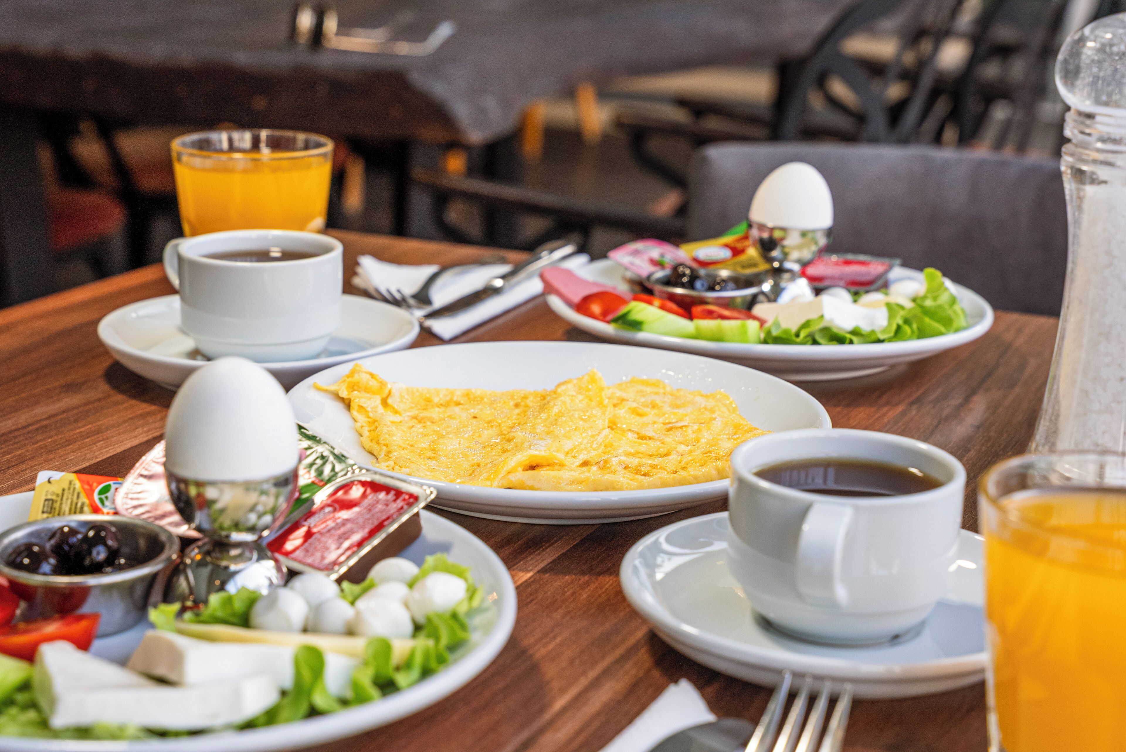 A breakfast setting with plates of eggs, toast, and salad, cups of coffee and orange juice, and condiments on a wooden table.
