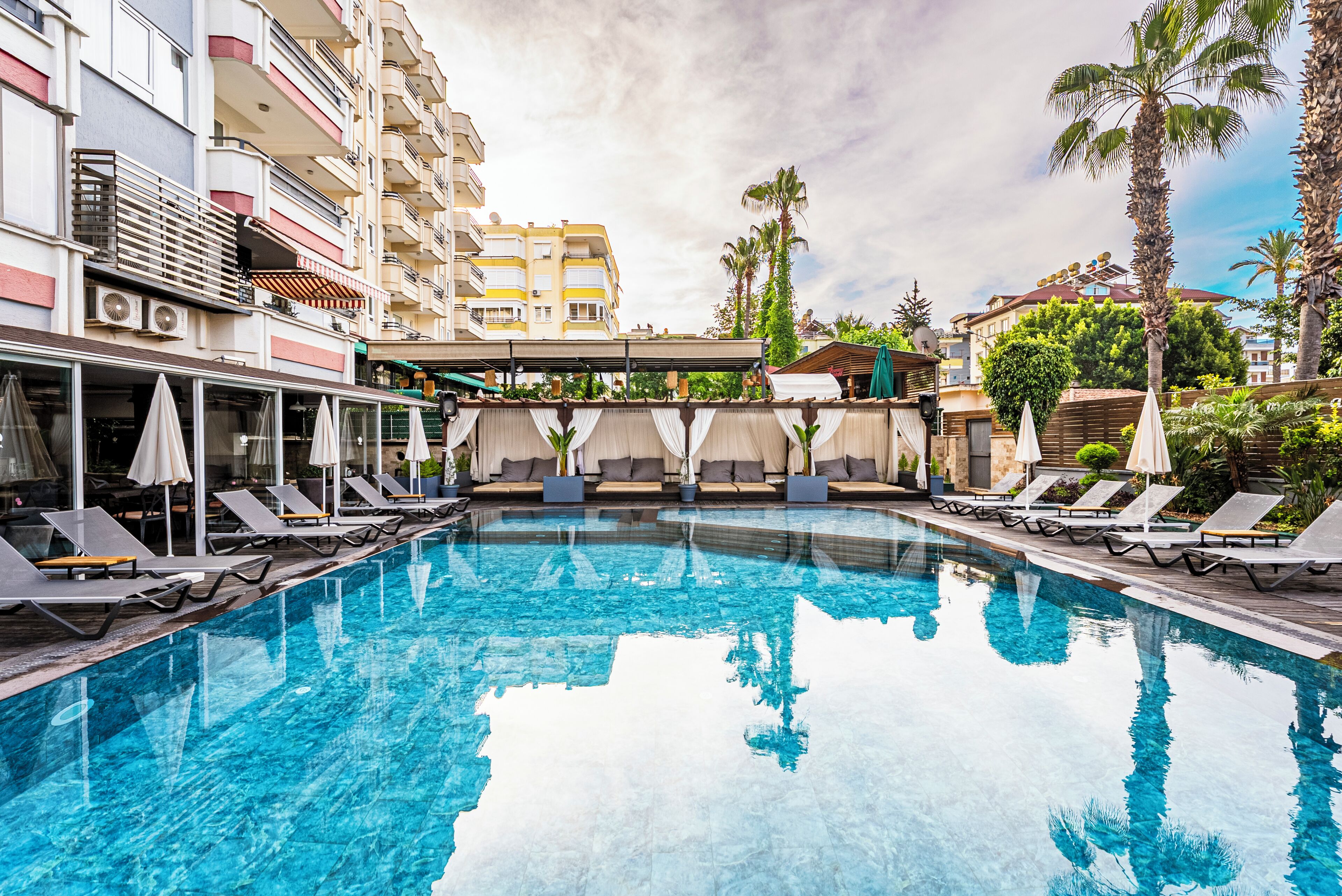 A swimming pool with lounge chairs and umbrellas, surrounded by multi-story buildings with balconies and outdoor seating areas.