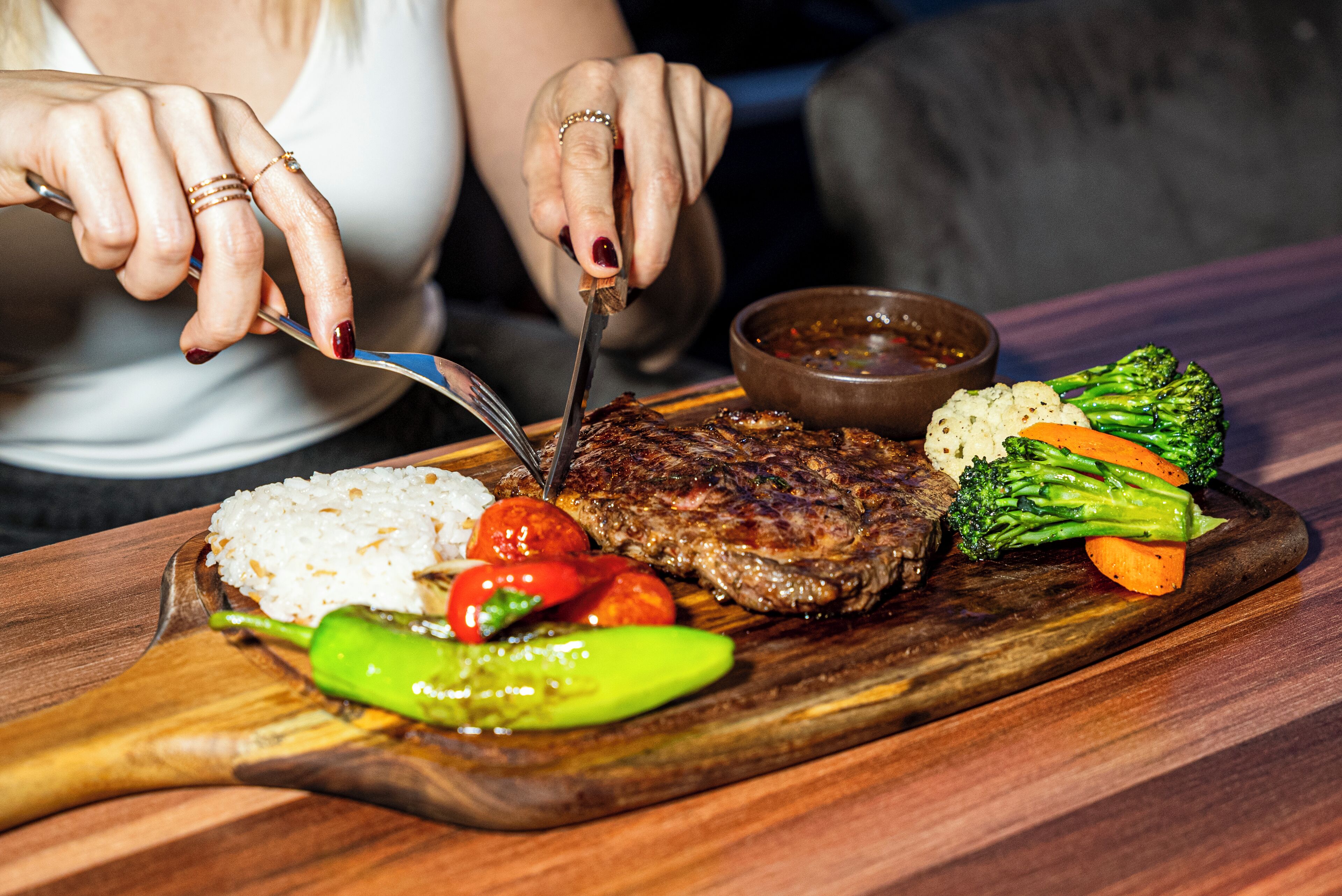 Une planche à découper en bois sur laquelle on voit un steak, du riz, des légumes et un ramequin de sauce.