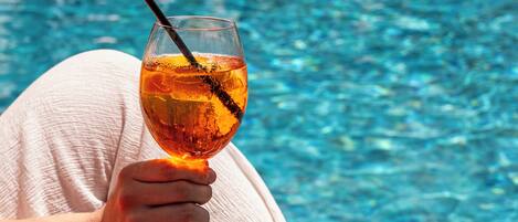Una persona sosteniendo un vaso con una bebida color ámbar junto a una piscina.