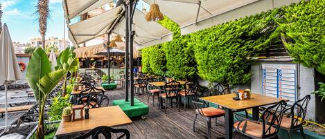 An outdoor café with wooden tables and chairs, surrounded by lush greenery and a thatched roof.