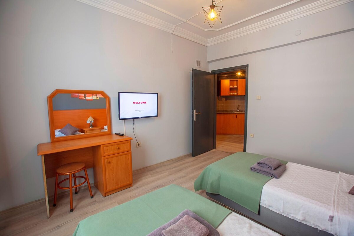 A hotel room with a desk, two beds, a TV displaying "WELCOME", and a kitchen area visible through an open door.