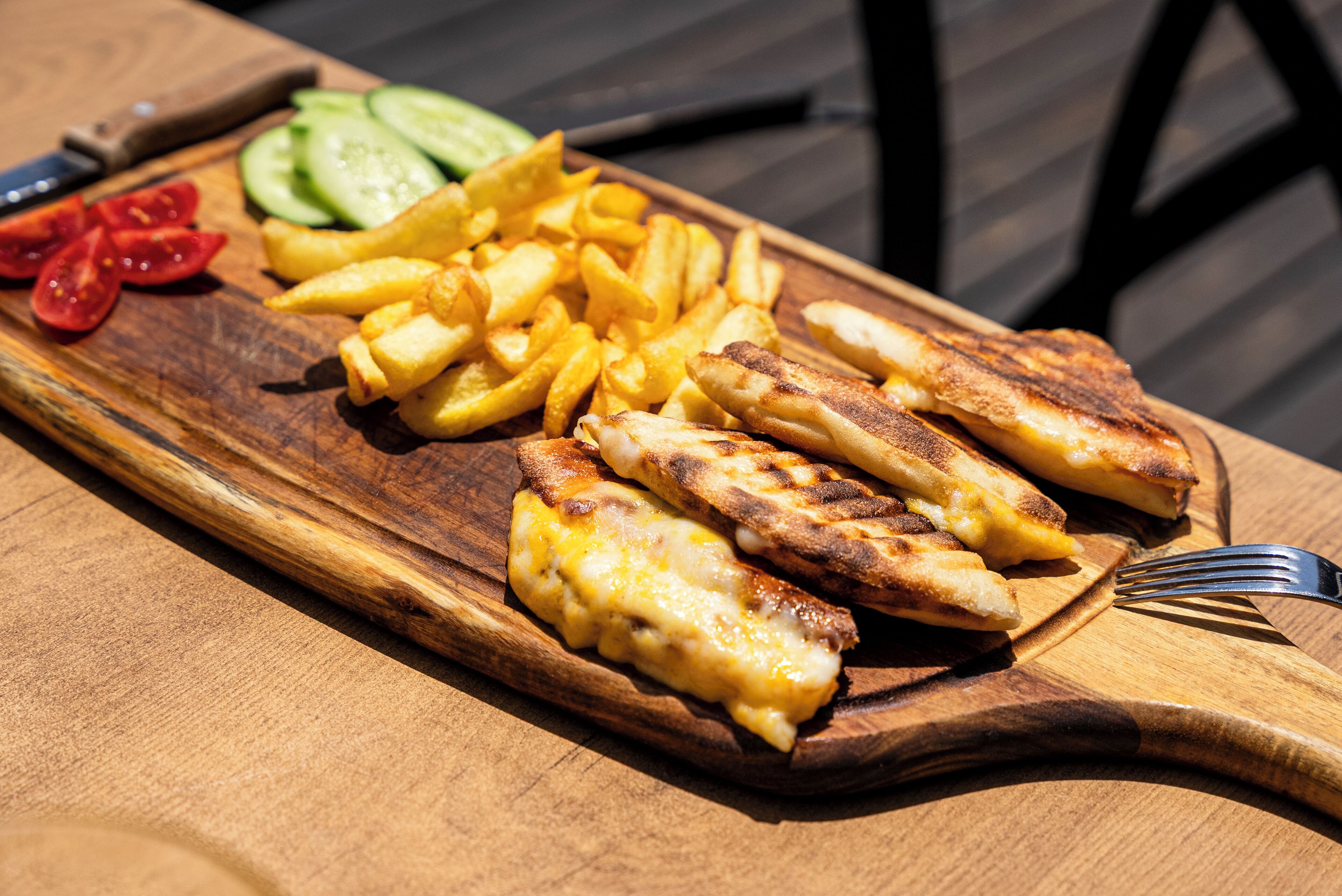 A wooden cutting board with grilled chicken, French fries, and sliced vegetables on a table.