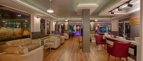 A spacious hotel lobby with a wooden floor, multiple seating areas including sofas and chairs, and a bar area with a counter and stools.