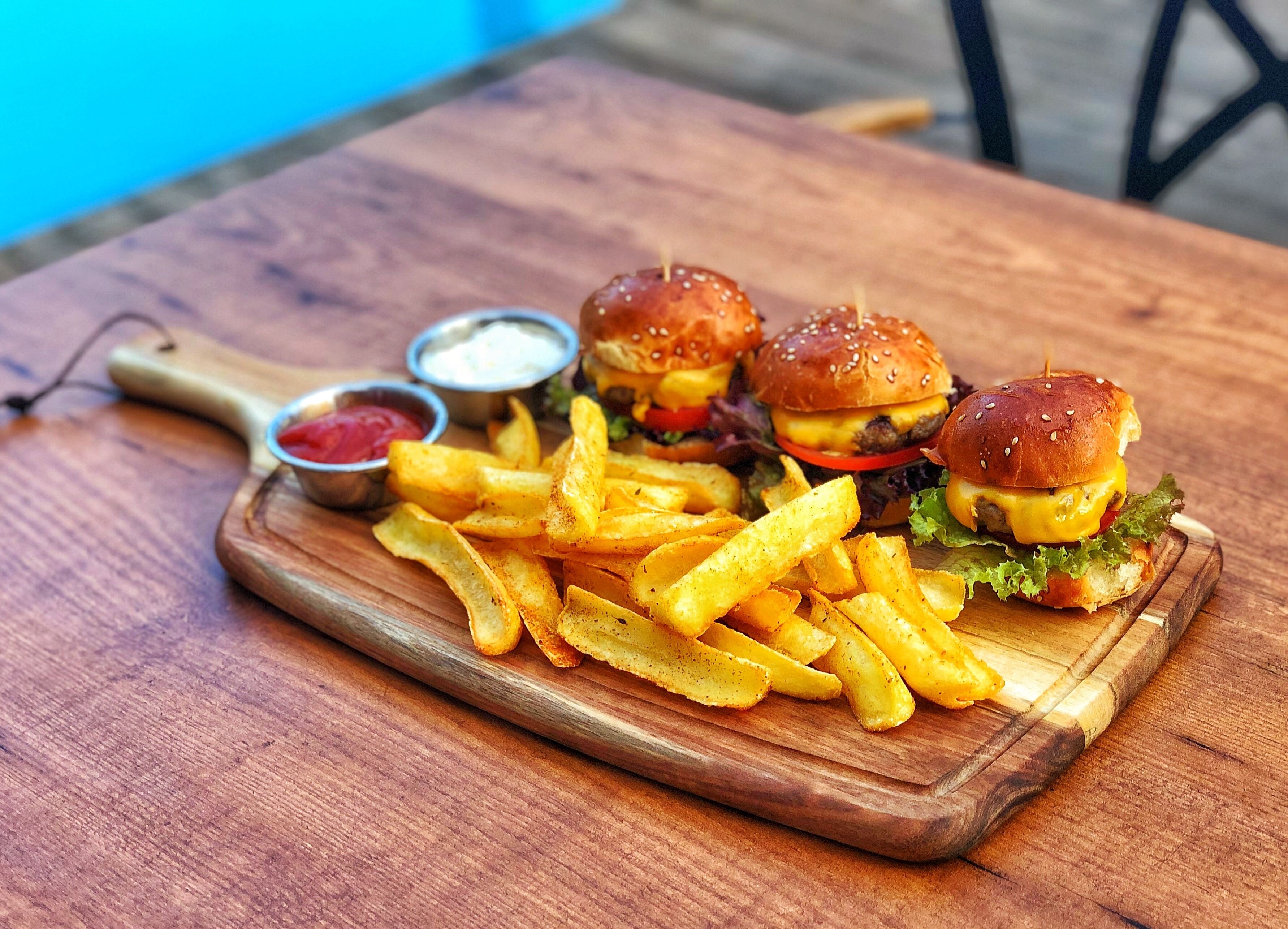 Una tabla de madera con tres hamburguesas y una porción de papas fritas, junto con dos recipientes pequeños de salsas para mojar.
