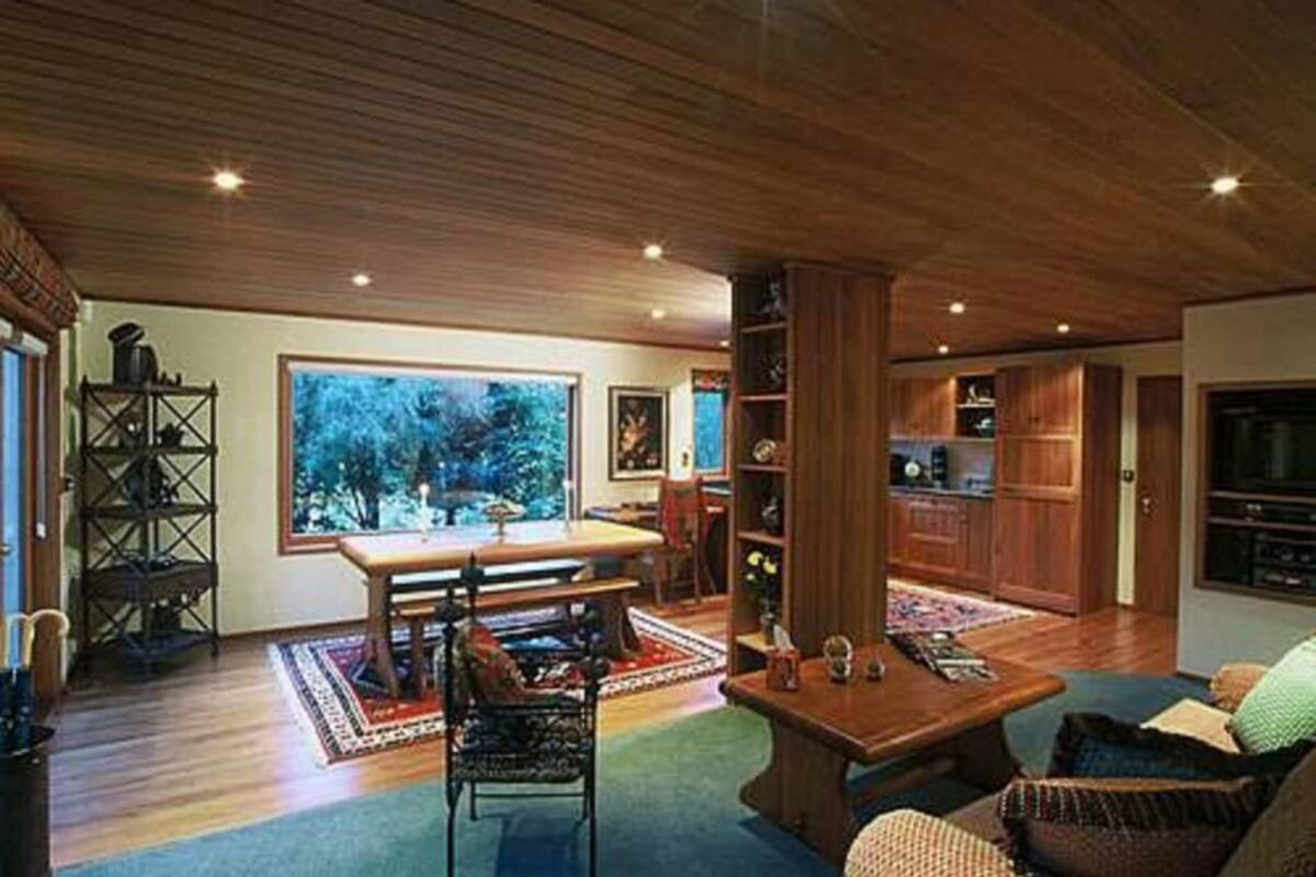 A spacious living room with a wooden ceiling, a large window offering a view of trees, a dining area with a table and chairs, and built-in wooden cabinets.