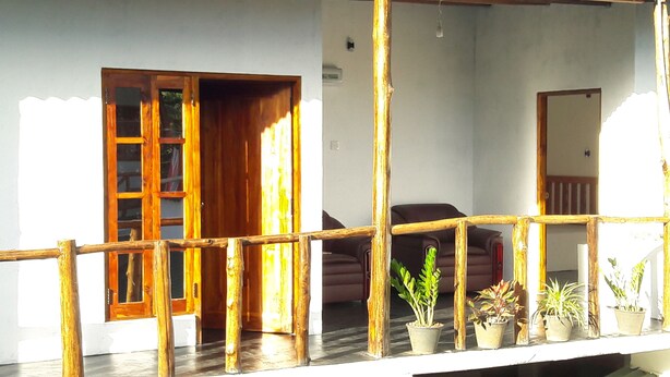 A wooden balcony with a railing, potted plants, and a view of the interior including a sofa and a door. - Eagle Safari Family Bungalow Udawalwe (Udawalawa)
