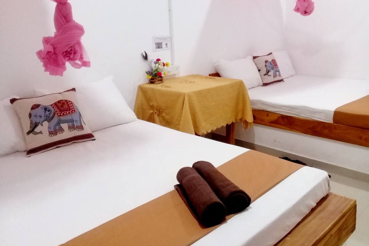 A hotel room with two beds, each with a white mattress and a wooden headboard. There is a small table with a yellow tablecloth and a bouquet of flowers.