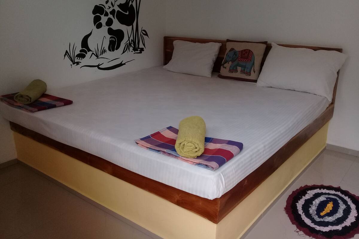 A bed with a white mattress, wooden headboard, and two pillows. A rolled towel and a colorful blanket are on the bed.