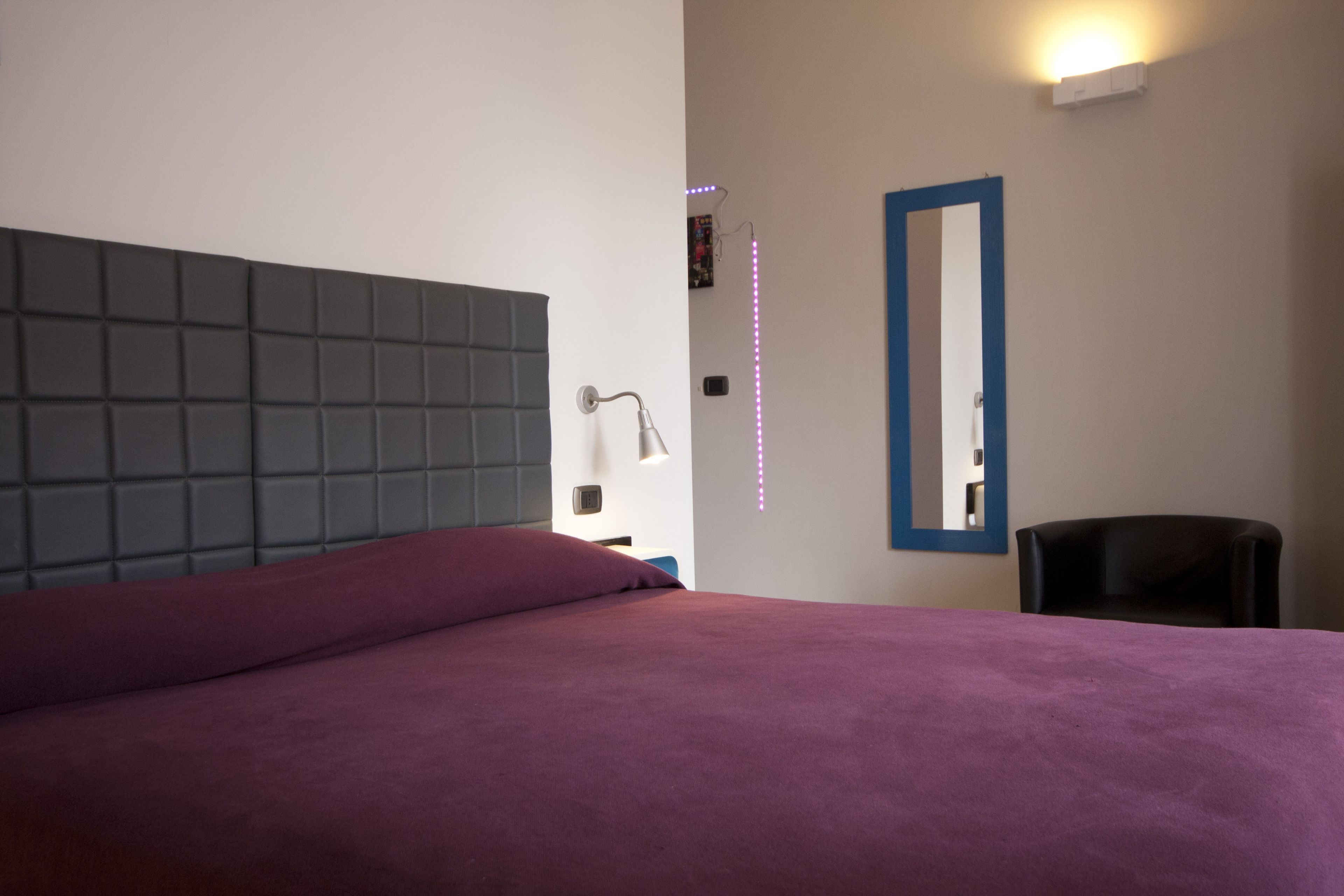 A bedroom with a large bed, a headboard, a mirror, a chair, and a wall-mounted light fixture.