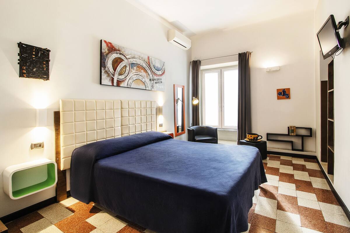 A hotel room with a large bed, a headboard, a wall-mounted TV, a painting, and a window with curtains.