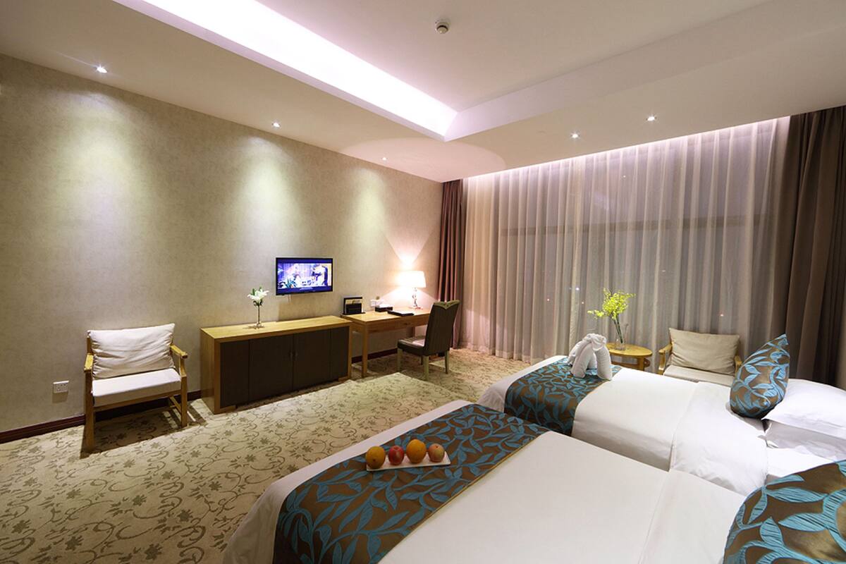Une chambre d’hôtel avec deux lits, un bureau avec une télévision, une chaise, une petite table avec un vase et une fenêtre avec des rideaux.
