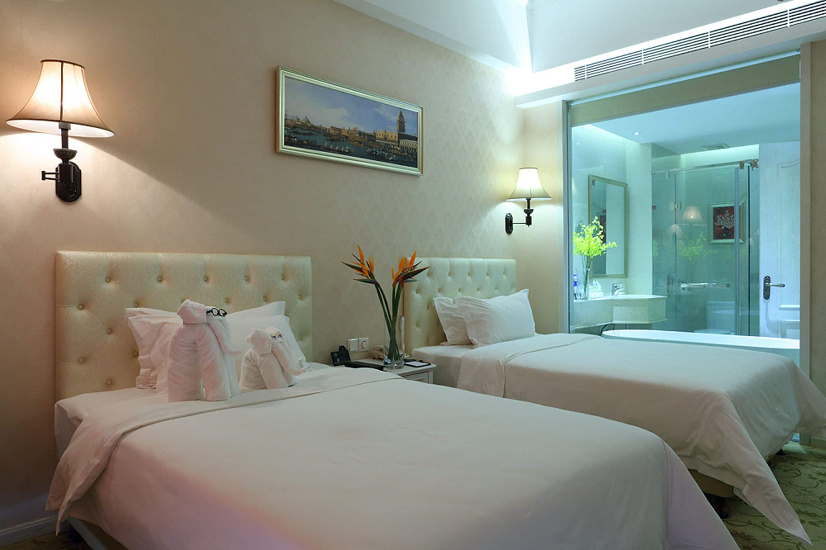 A hotel room with two beds, a large window, and a bathroom visible through the glass door.