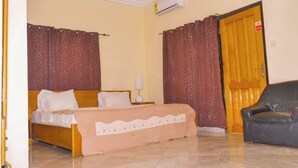A hotel room with a bed, a nightstand, a lamp, a chair, and a door. - Jenos Hotels (Accra)
