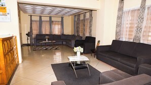 A living room with a sectional sofa, a small table with a vase, and a fan. - Jenos Hotels (Accra)