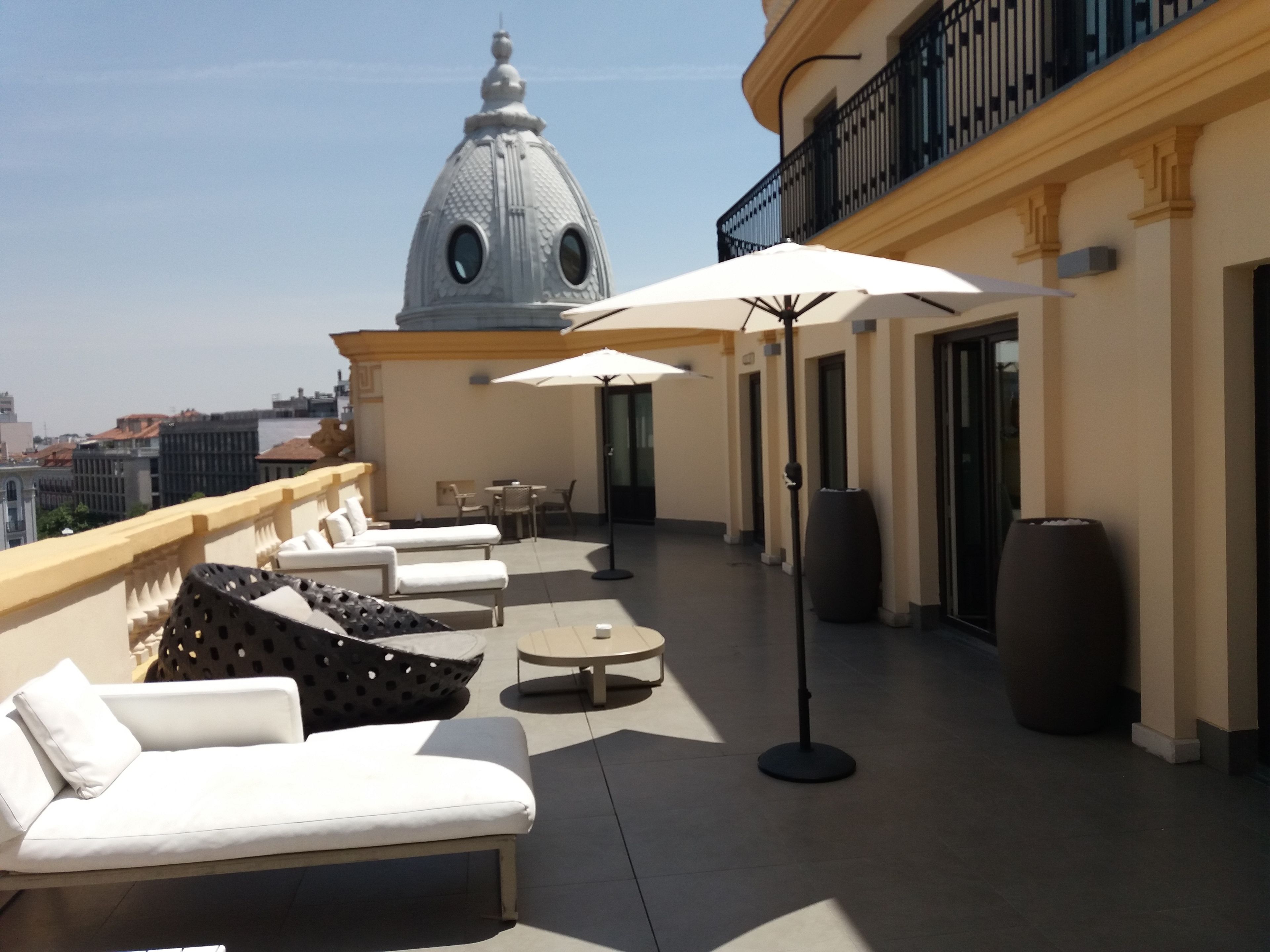 A rooftop terrace with white lounge chairs, a table, and umbrellas. A large dome structure is visible in the background.