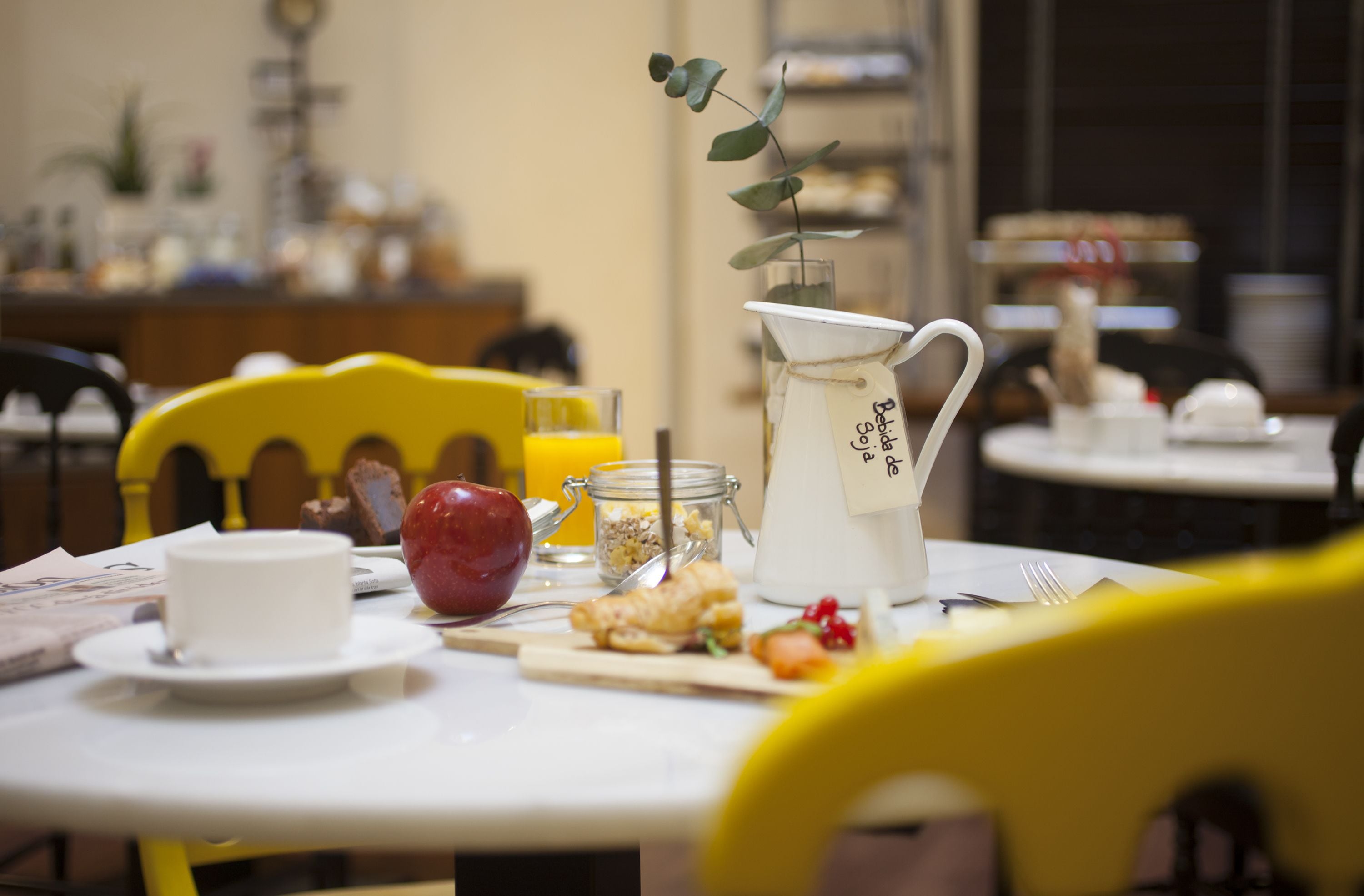 A breakfast table with a white pitcher, a red apple, a glass of orange juice, and a small bowl of cereal.