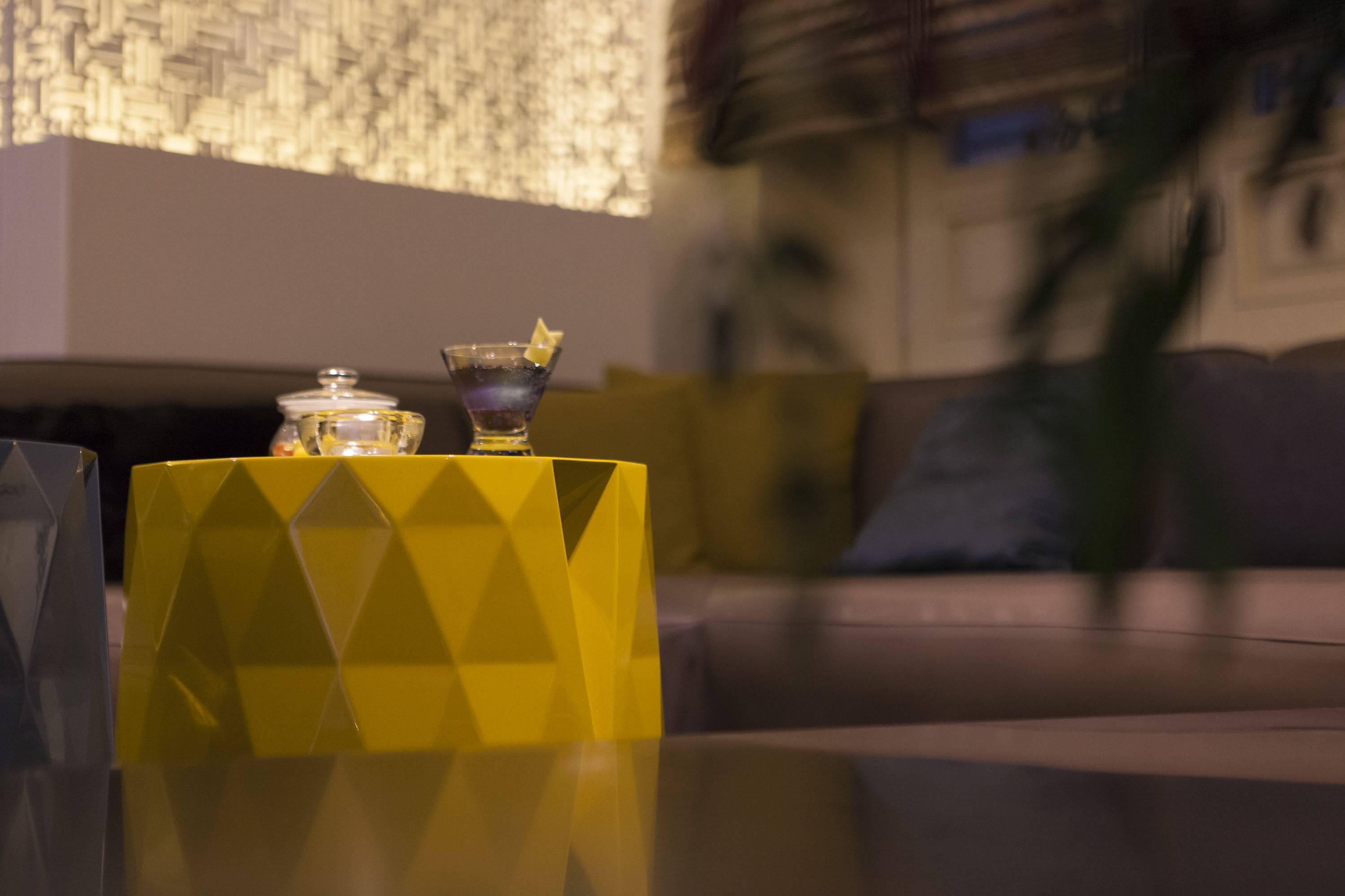 A yellow geometric-patterned box on a reflective surface, a glass container with a lid, and a blurred background with a couch and a plant.