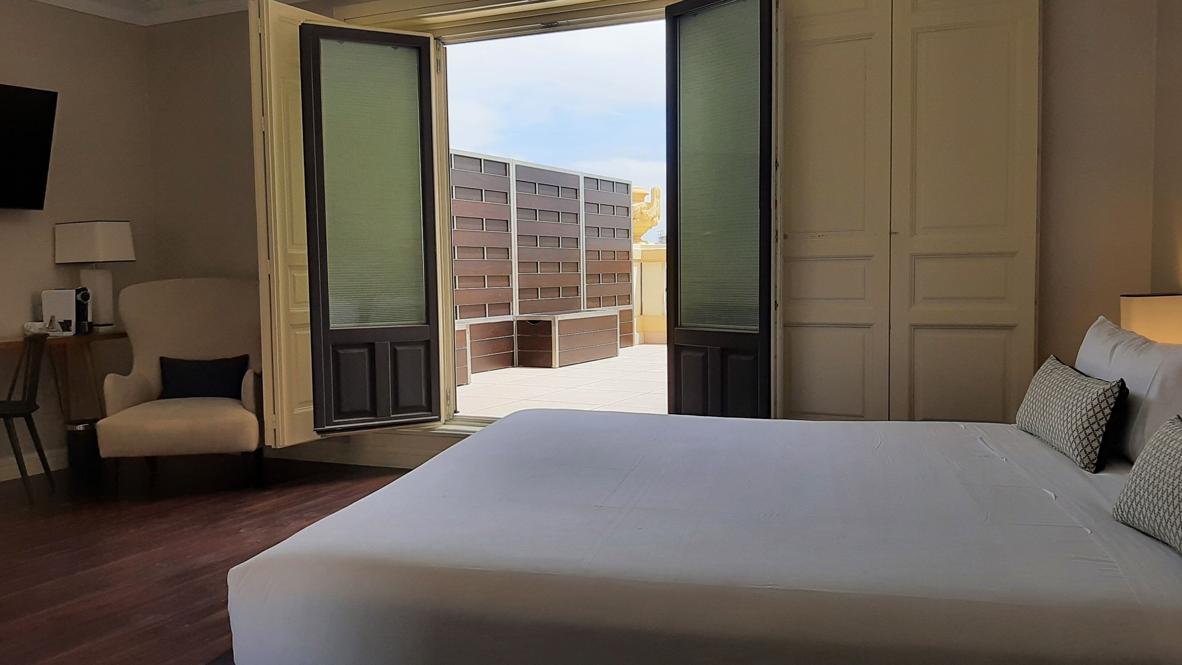 Une chambre d’hôtel avec un grand lit, une chaise, un bureau avec une lampe et une vue sur un bâtiment à l’extérieur.