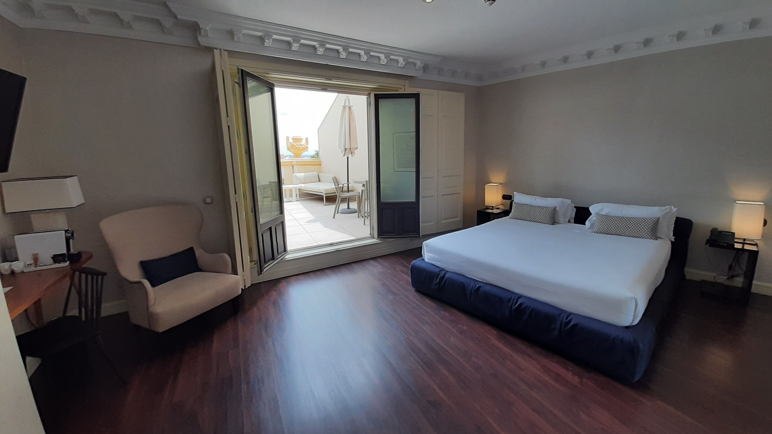 Una habitación de hotel con una cama grande, una silla, un escritorio con lámpara y un ventanal corredero que da a un balcón.