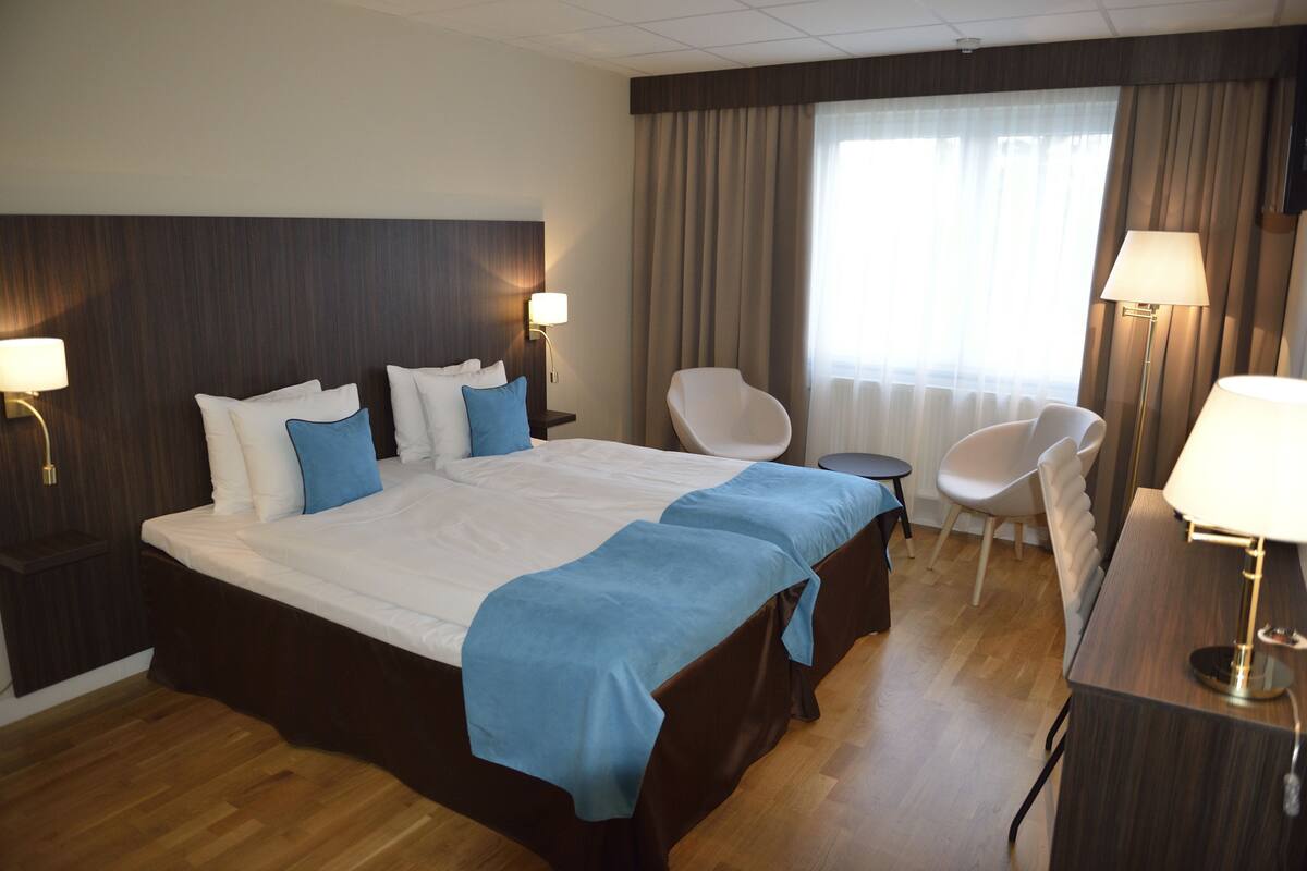 A hotel room with a large bed, two bedside tables with lamps, a chair, a desk with a lamp, and a window with curtains.