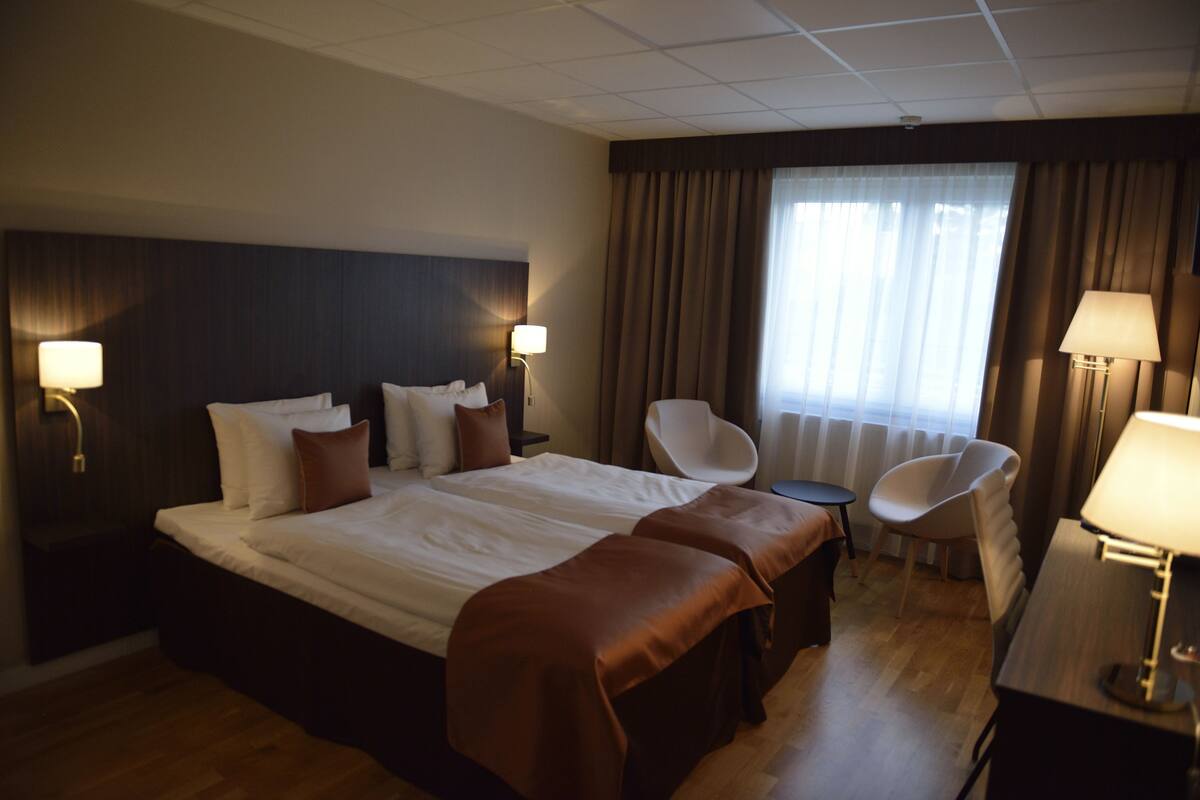 A hotel room with a large bed, two bedside tables, a desk, a chair, and a window with curtains.