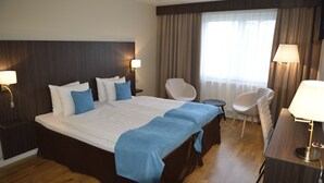 A hotel room with a large bed, two bedside tables with lamps, a chair, a desk with a lamp, and a window with curtains. - Hotell Nova (Karlstad)