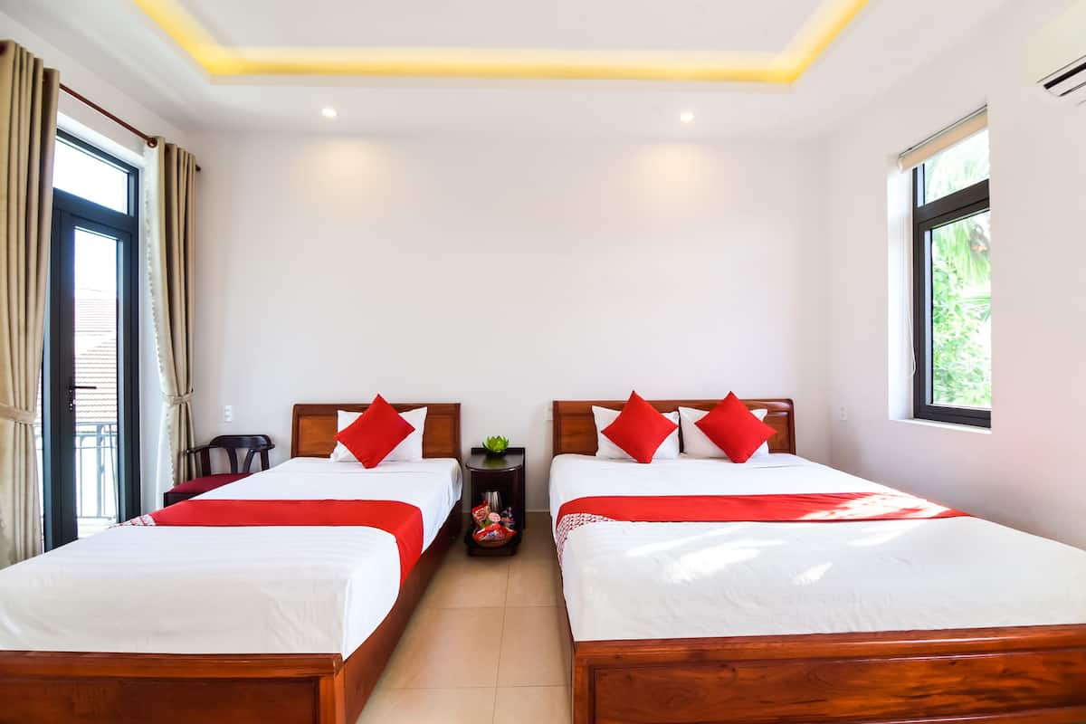 Habitación de hotel con dos camas de madera, cada una con ropa de cama roja y blanca, una mesita con una planta y una ventana que ofrece vistas al verdor.