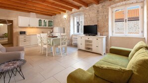 A cozy living room with a green sofa, a dining area with white chairs and a table, a kitchen with white cabinets, and a stone wall. - Vila Mama (Omis)