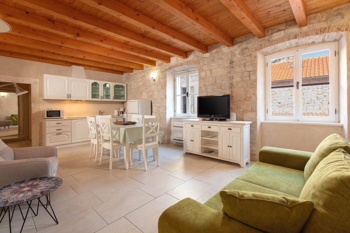 A cozy living room with a green sofa, a dining area with white chairs and a table, a kitchen with white cabinets, and a stone wall.