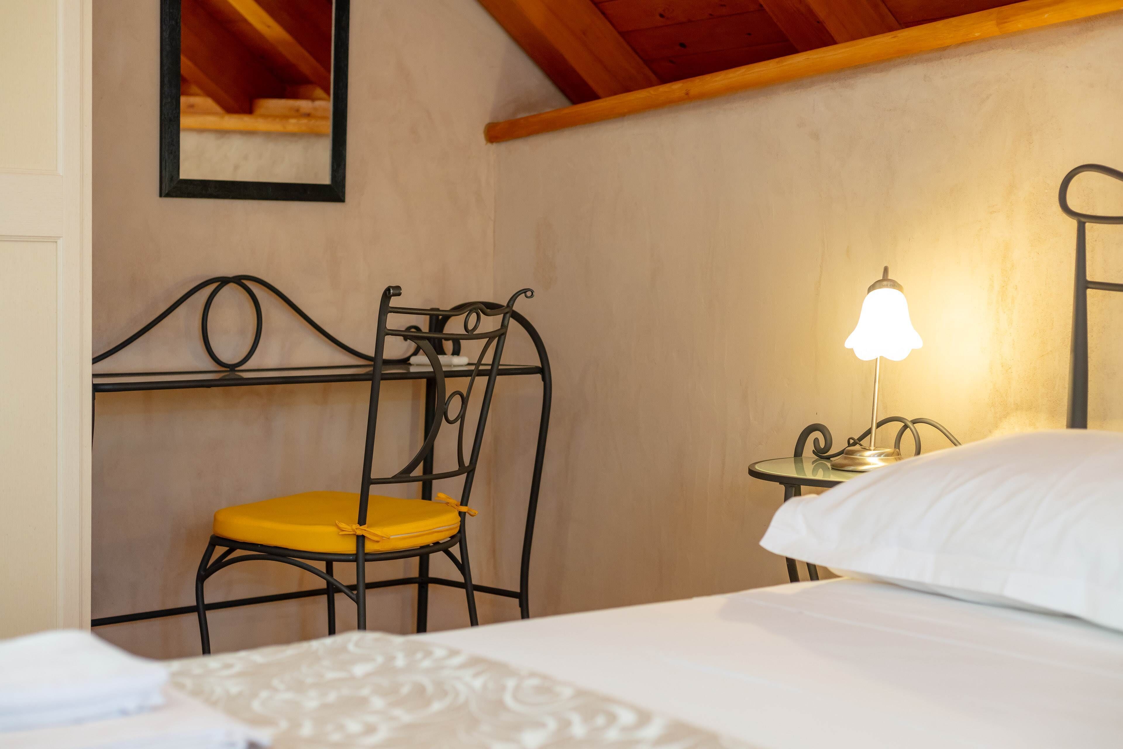 Un dormitorio con cama, mesita de noche con lámpara, una silla y espejo en la pared.