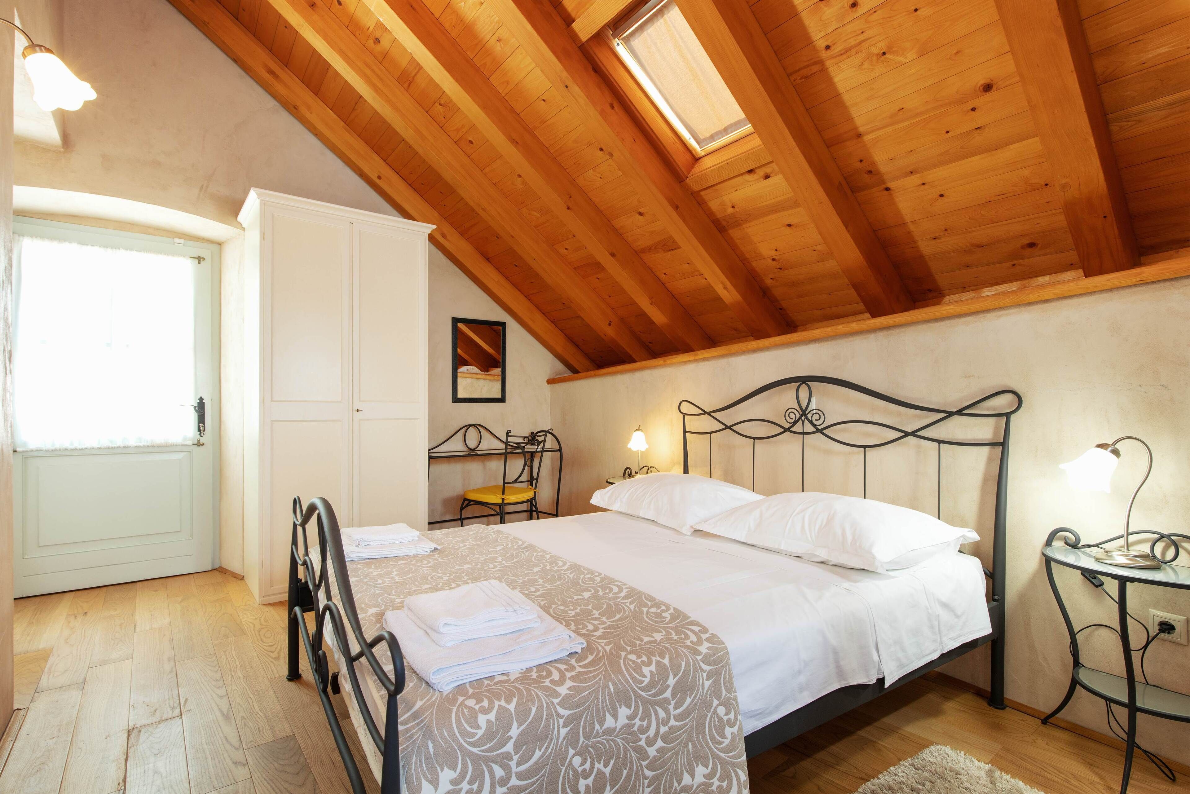 Un dormitorio con techo de madera, una cama con sábanas blancas, una mesita de noche con una lámpara y una silla.