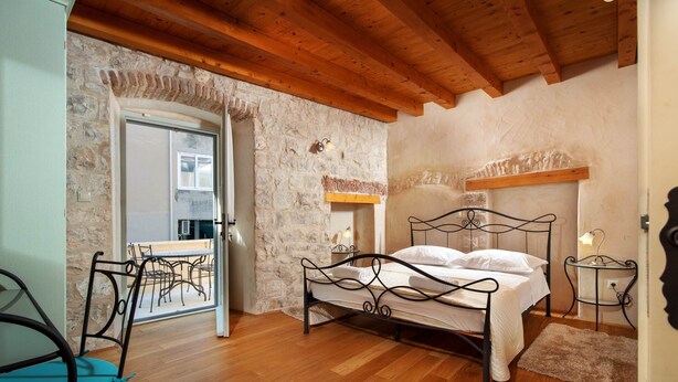 A bedroom with a stone wall, wooden floor, a bed with a metal frame, a chair, and a table with a lamp. - Vila Mama (Omis)