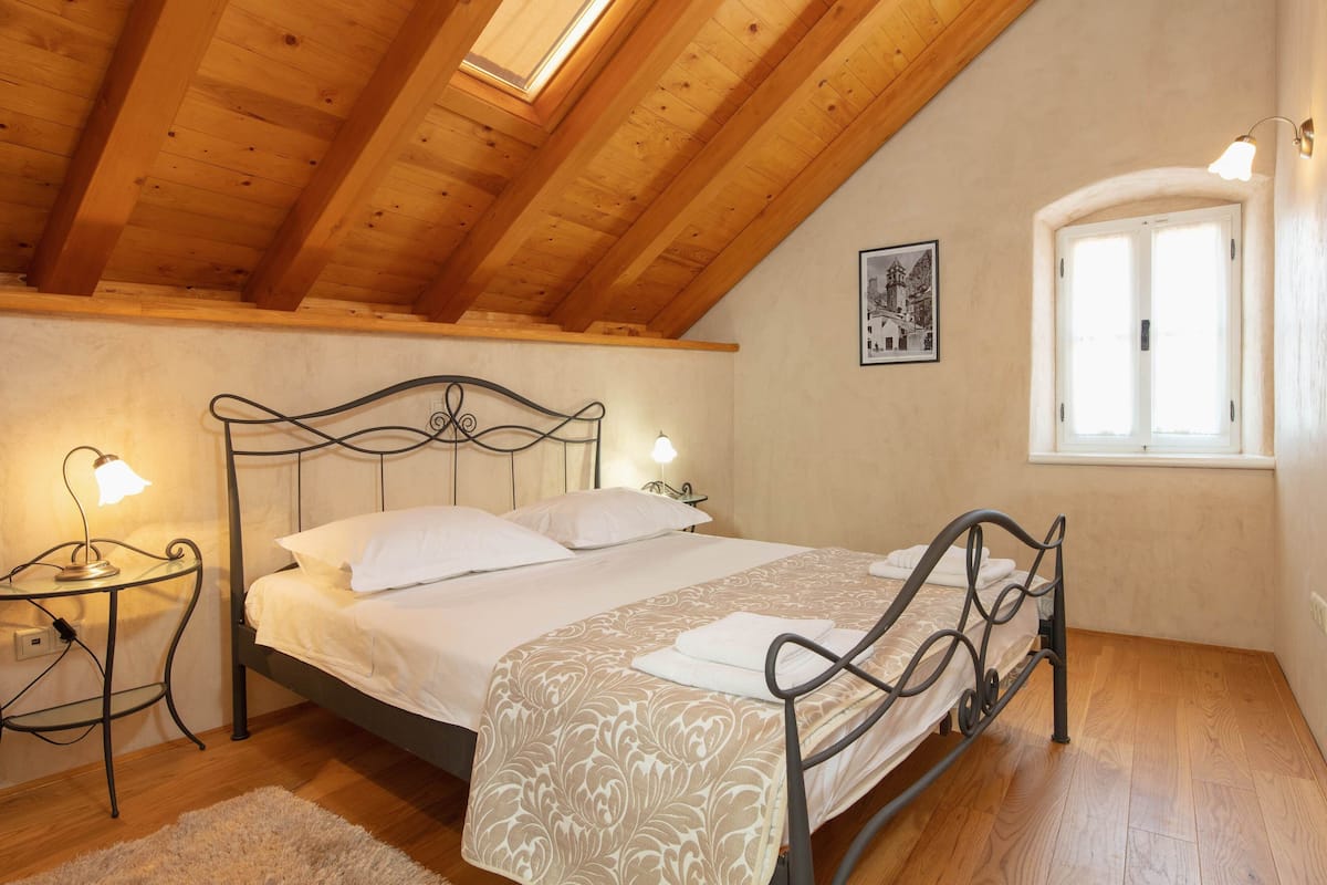 A bedroom with a wooden ceiling, a bed with a patterned bedspread, two bedside lamps, a window with white shutters, and a framed picture on the wall.