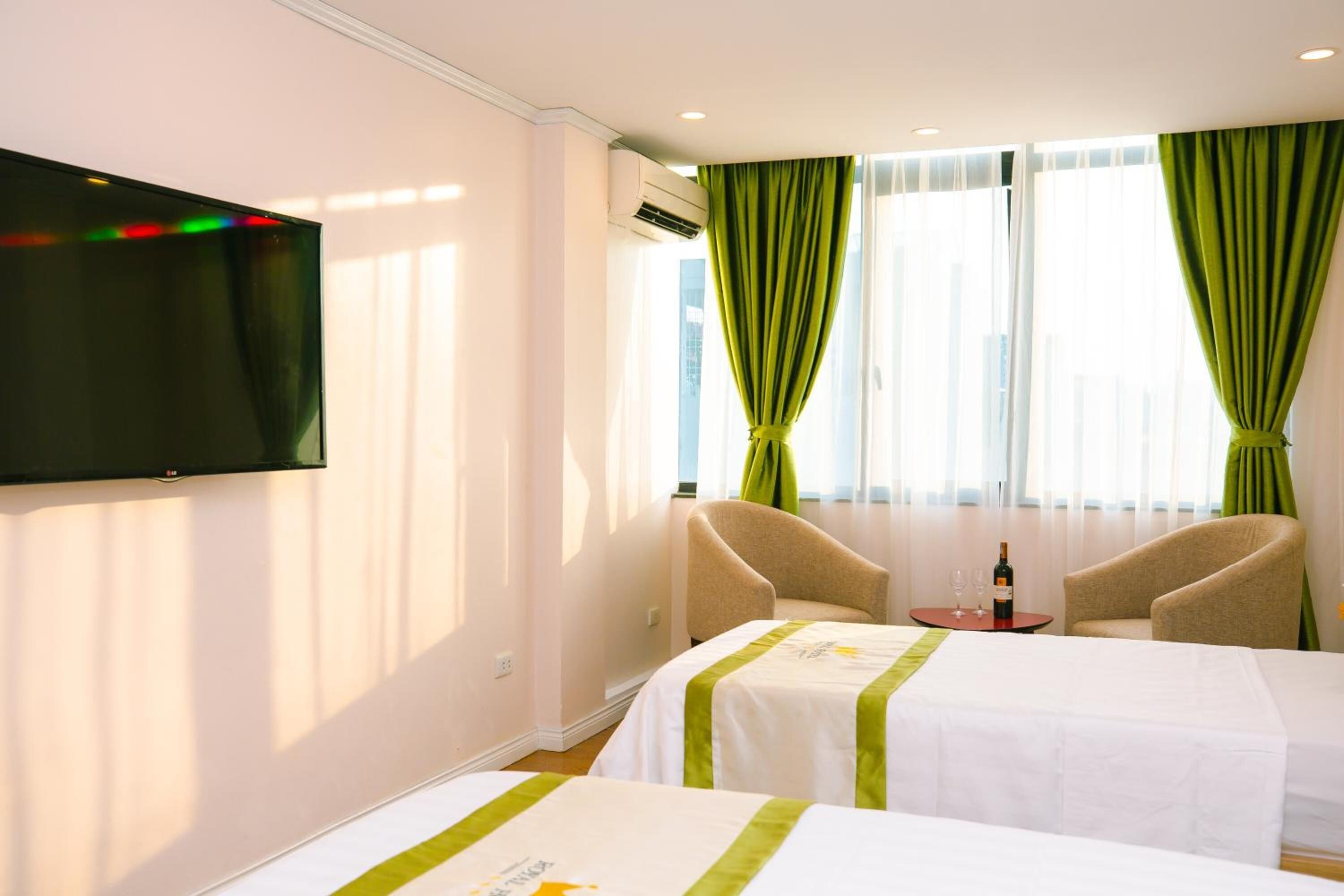 A hotel room with two beds, a TV mounted on the wall, a small table with a bottle and glasses, and a window with green curtains.