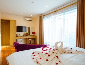 A hotel room with a bed adorned with rose petals and a heart-shaped arrangement, a wooden desk with a television, a minibar, and a purple chair. - Royal Hotel (Hanoi)