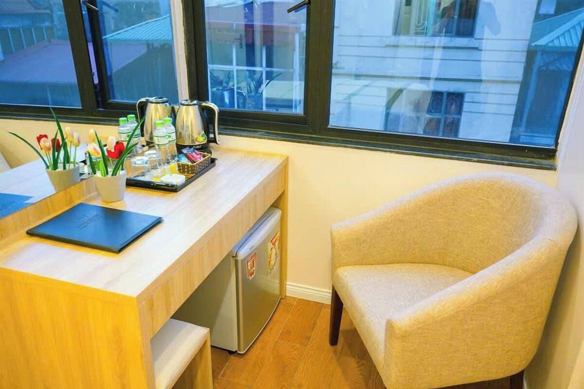 A small, tidy room with a wooden desk, a chair, a window, and a refrigerator.