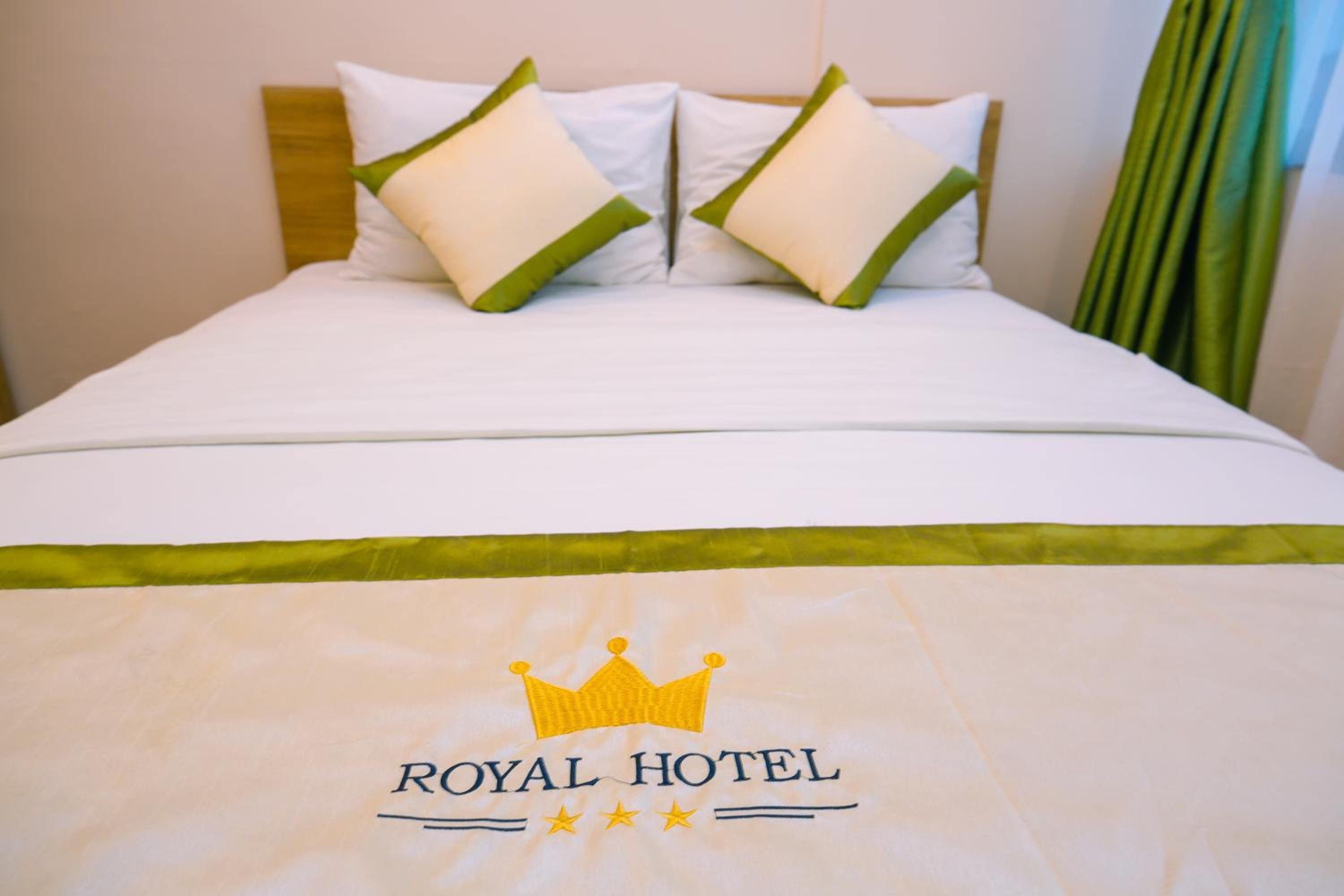 Uma cama de hotel com roupa de cama branca, almofadas e colcha verdes, com o logotipo do Royal Hotel.