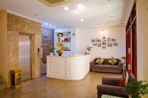 A hotel lobby with a reception desk, a sofa, a coffee table, a small plant, and a wall with framed pictures. - Royal Hotel (Hanoi)