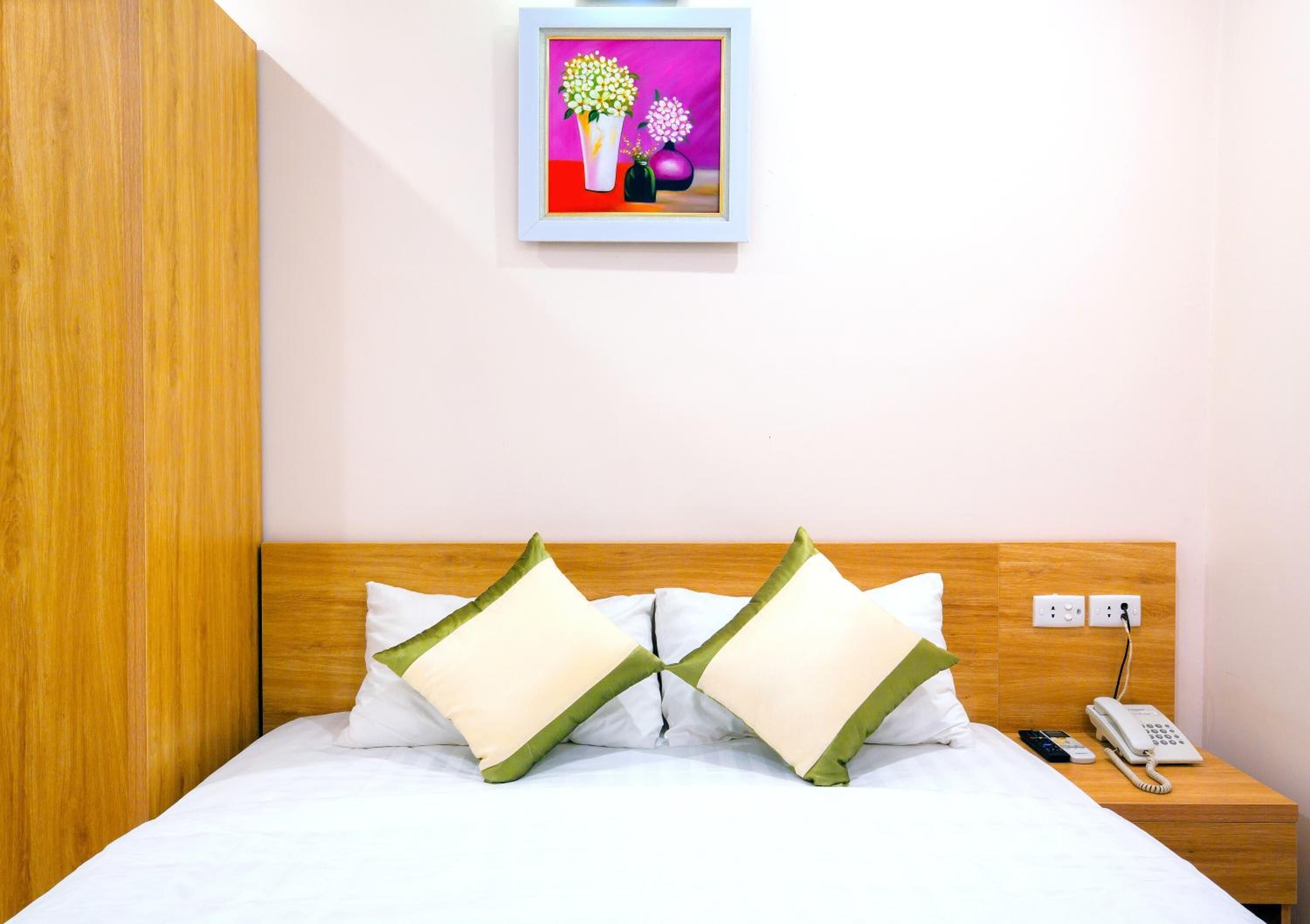 A hotel room with a wooden headboard, a bed with white bedding, two green and white pillows, a bedside table with a phone, and a framed picture on the wall.