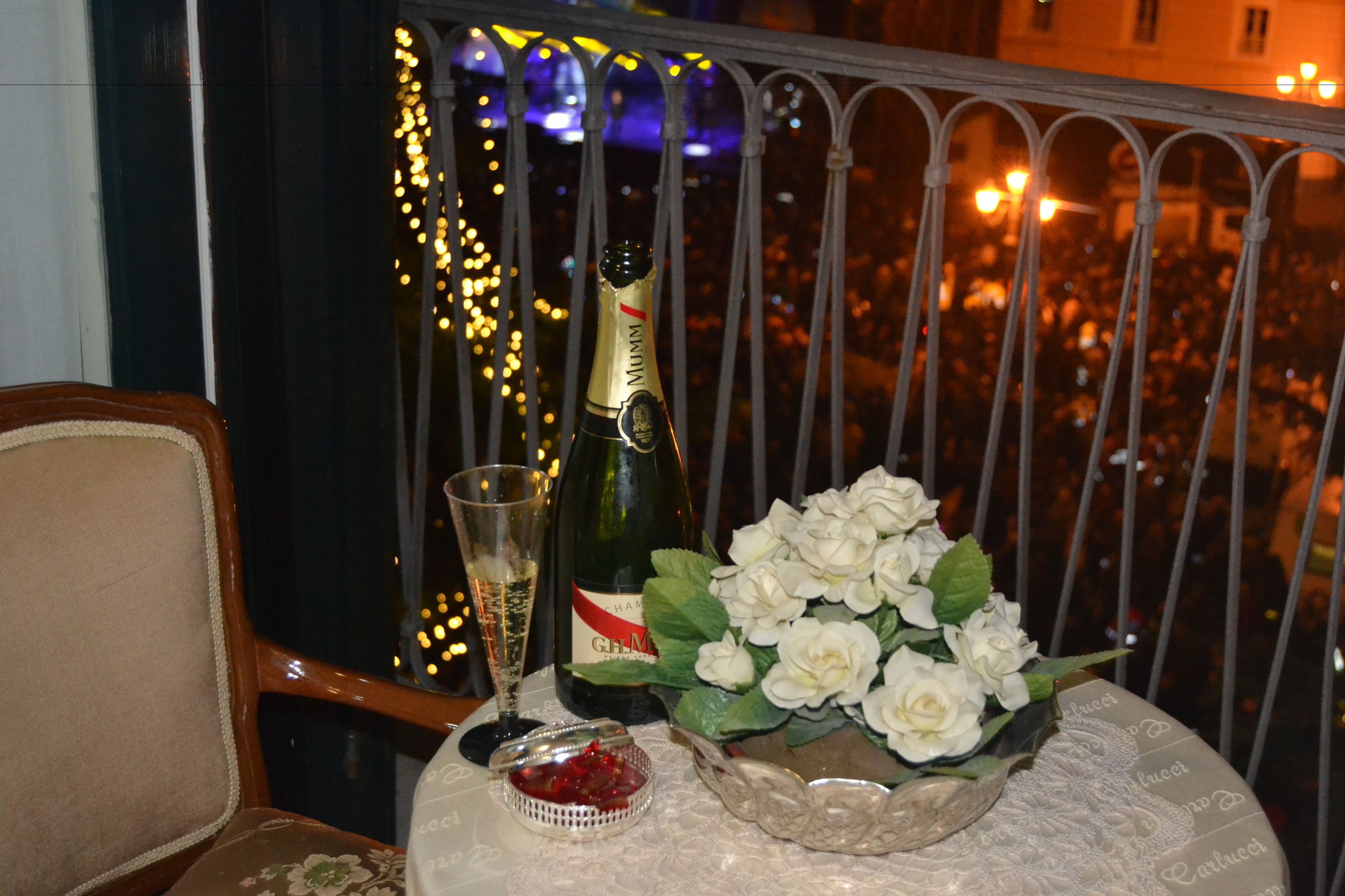 Un balcone con un tavolo apparecchiato: una bottiglia di champagne, un bicchiere, una ciotola di rose bianche e un piccolo recipiente con bacche rosse.