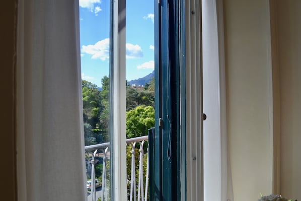 Vista a través de una ventana con cortina blanca, mostrando un balcón con barandilla, una calle con coches aparcados y vegetación al fondo.