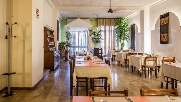 A restaurant with multiple tables and chairs, a long counter with a cash register, and a ceiling fan. - Hotel Villa Rosa (Sarroch)