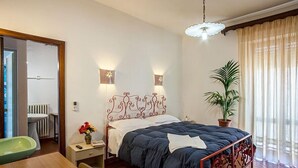 A bedroom with a wooden bed frame, two bedside lamps, a potted plant, and a view of a bathroom through an open door. - Hotel Villa Rosa (Sarroch)
