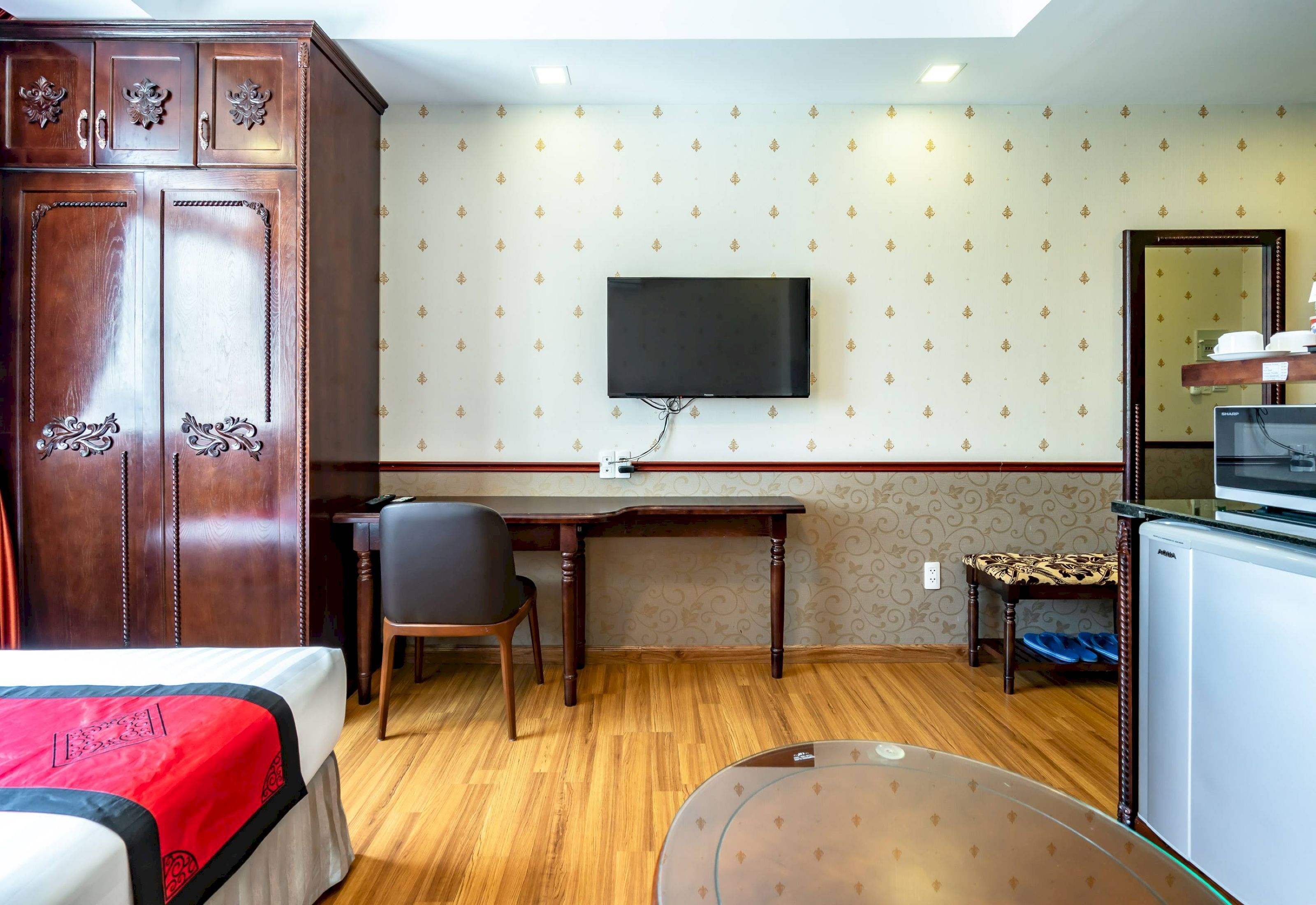 A hotel room with a wooden wardrobe, a desk with a chair, a television mounted on the wall, a bed with a red and white bedspread, and a small table with a glass top.
