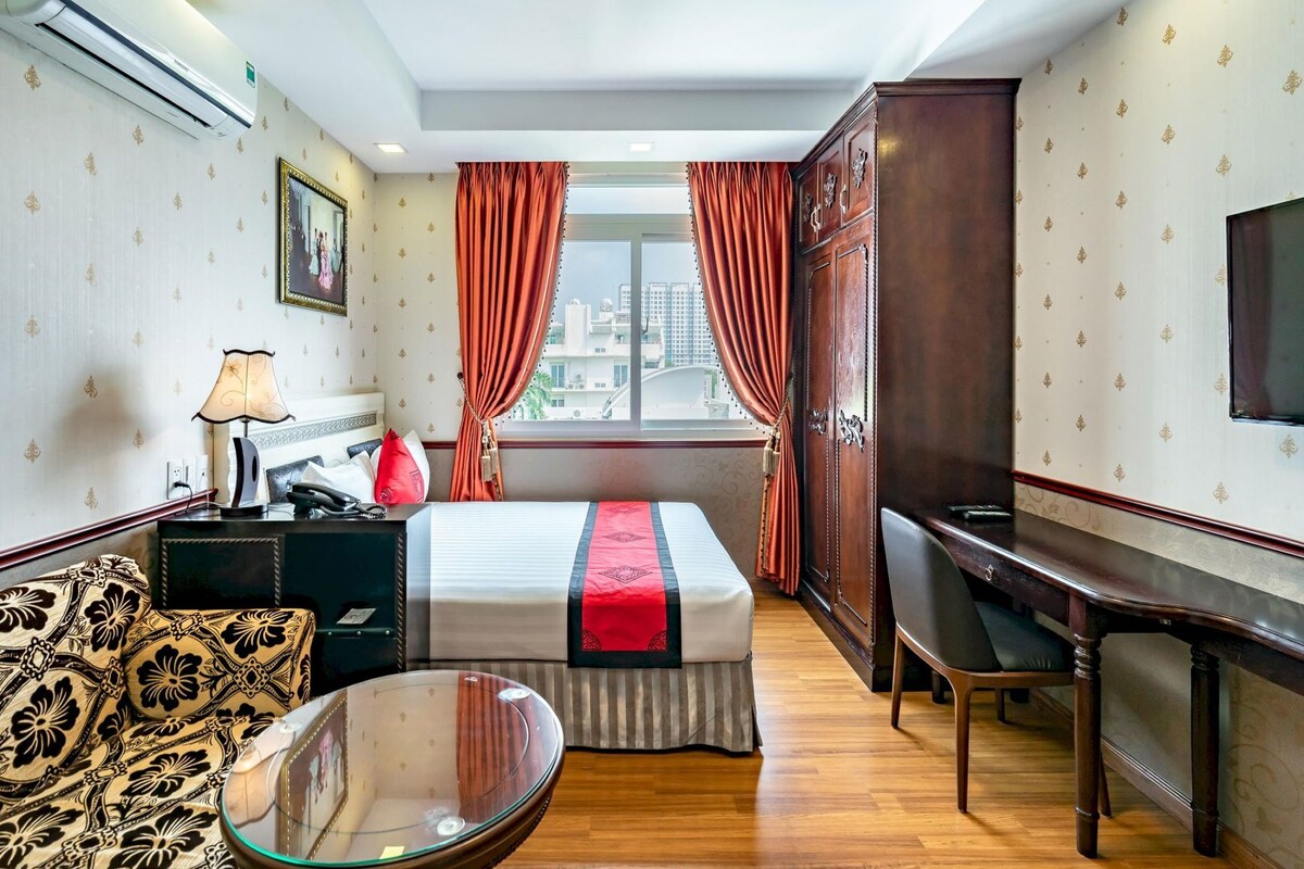 Une chambre d’hôtel avec un lit, un bureau, une chaise, une table à plateau en verre et une vue sur la ville par la fenêtre.