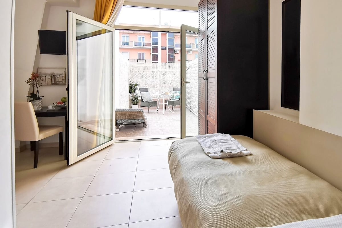 A bedroom with a bed, a desk, and a chair. There is a sliding glass door leading to an outdoor area with a table and chairs.